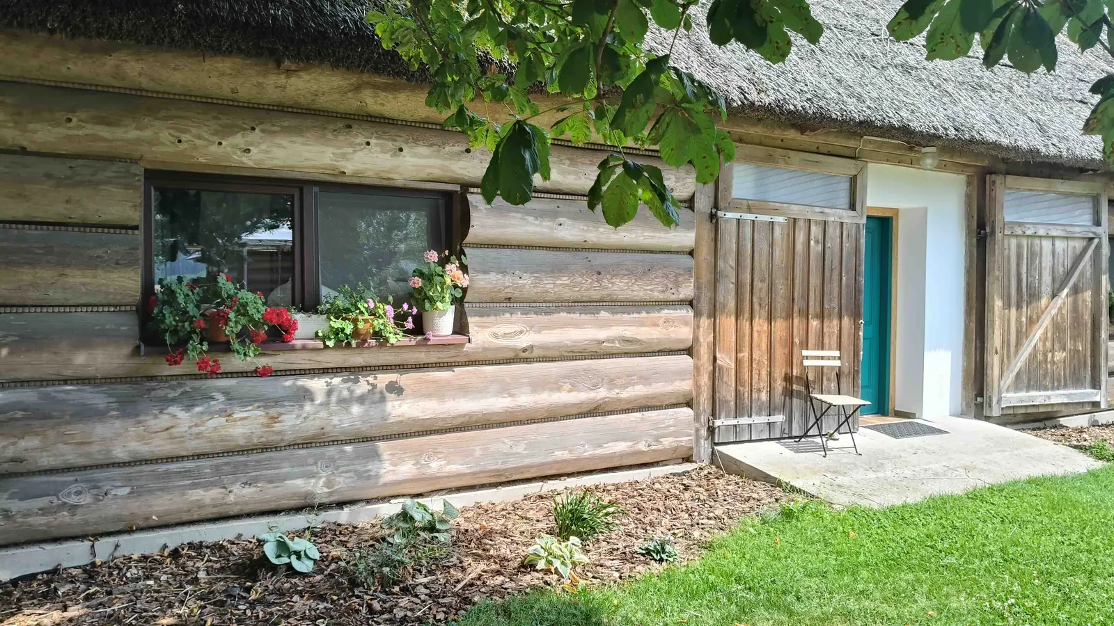 Ściana z bali, okno z pelargoniami i turkusowe drzwi Stodoły